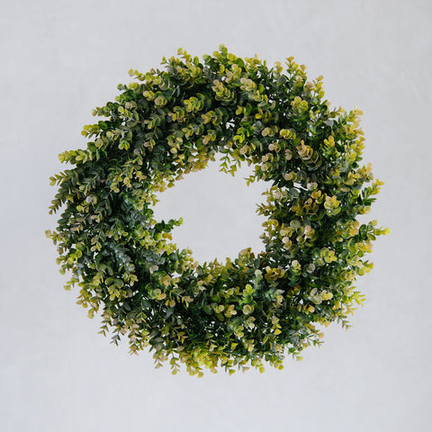 An artificial boxwood wreath with dense green and yellow-green foliage, centered against a plain light gray background.