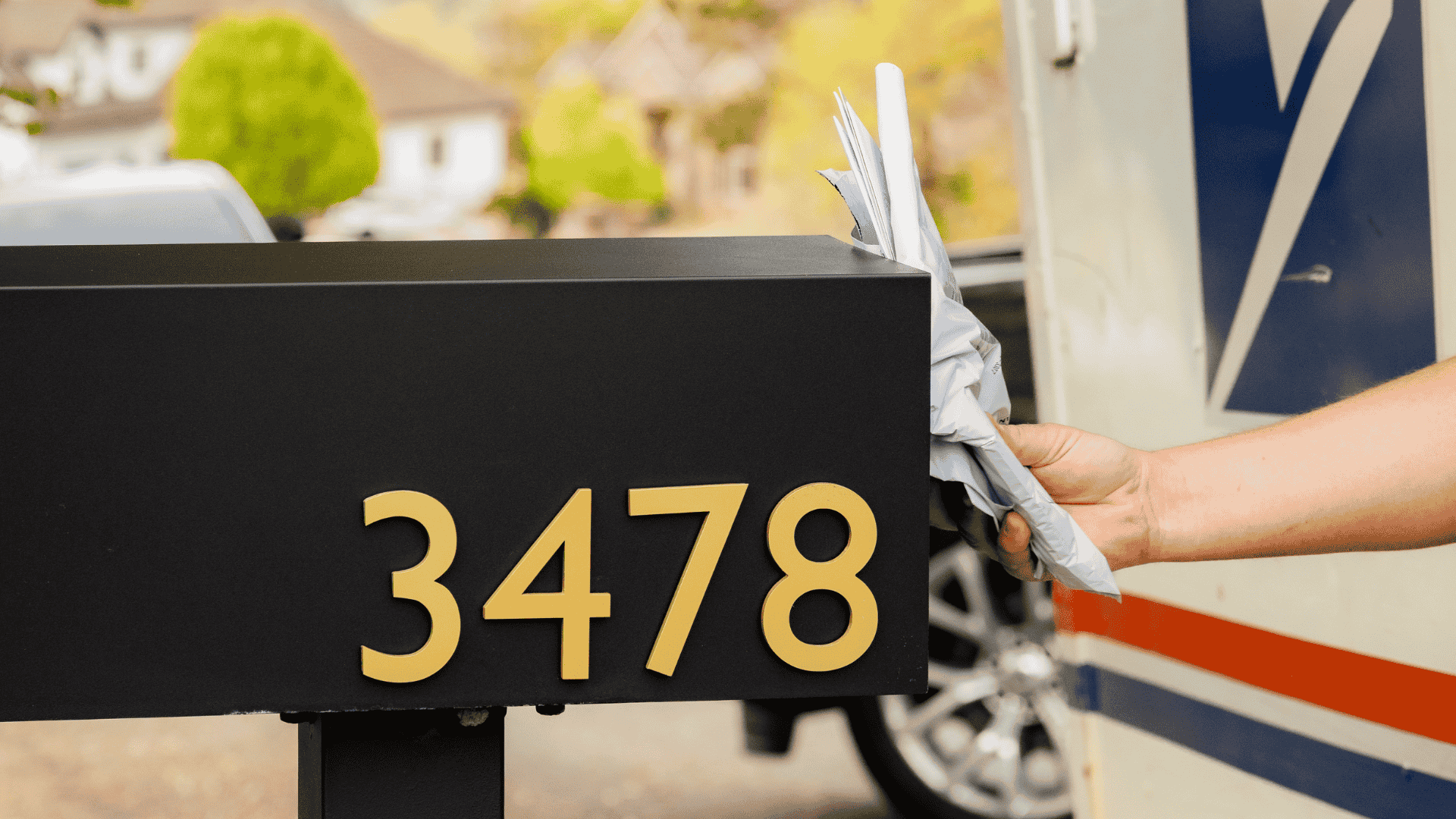 Rear-access mailbox with gold address numbers 3478, hand placing mail into it, blurred background of a residential area.