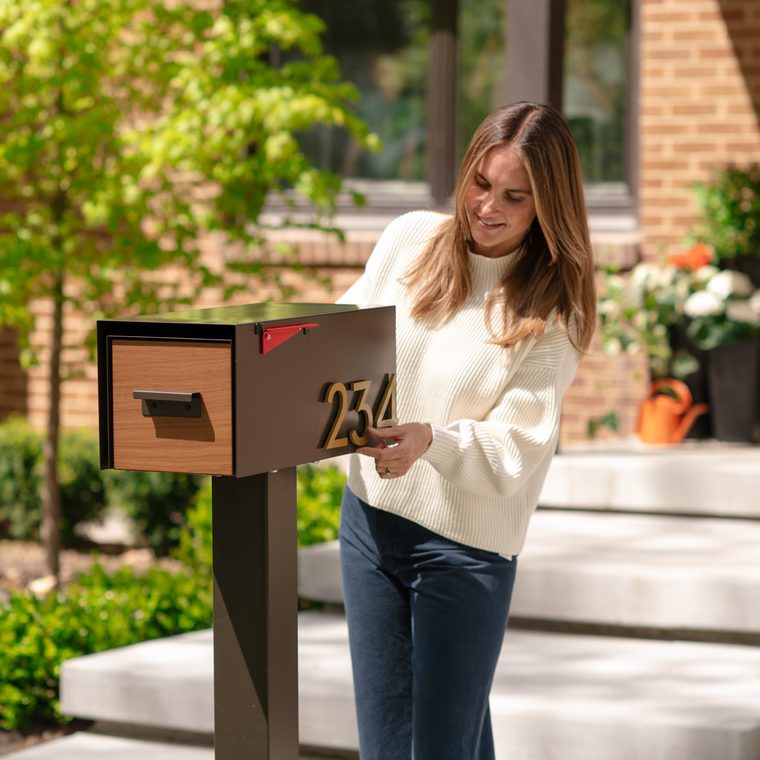 A homeowner opening The Malone post-mounted mailbox with a wood door and house numbers 234, in front of a brick home